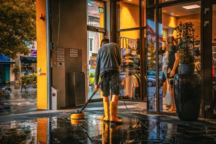 Un hombre limpia el piso de la entrada al interior de una tienda en un día lluvioso, reflejando la vida urbana.